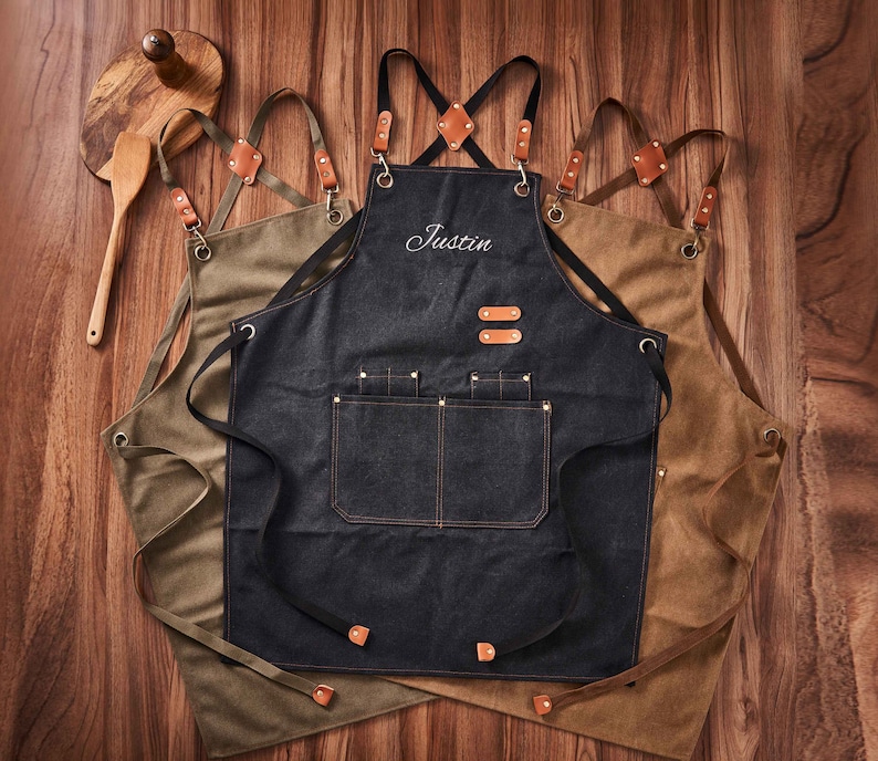 May include: Three brown canvas aprons with adjustable straps. The centre apron is black denim with a large pocket and the name "Justin" embroidered on the front.