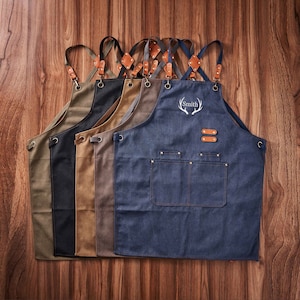 May include: Five denim aprons with adjustable straps and leather accents. One apron has the name "Smith" embroidered on it with a stag antler design.