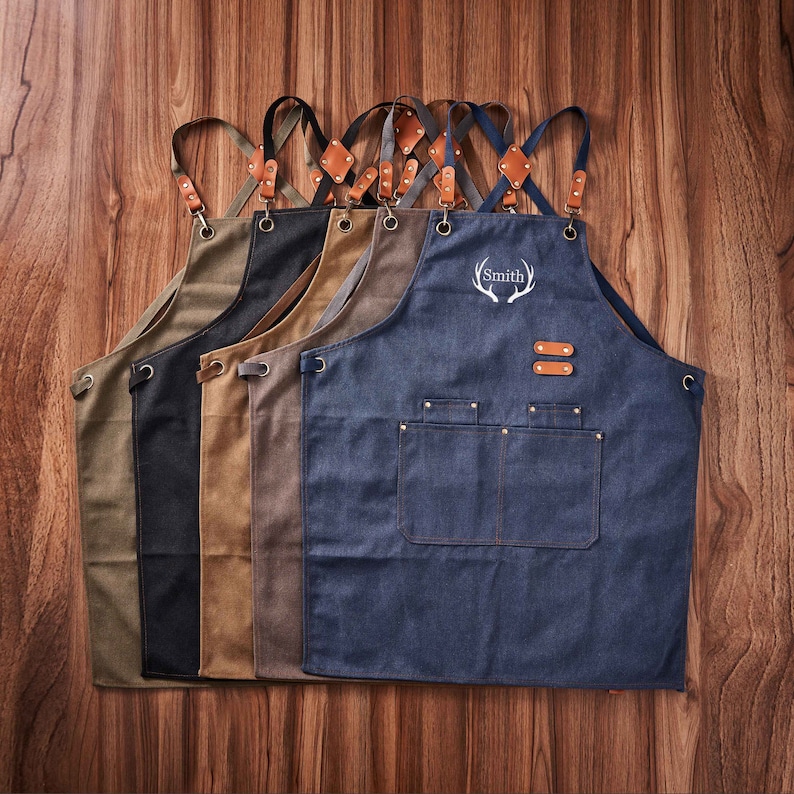 May include: Five denim aprons with adjustable straps and leather accents. One apron has the name "Smith" embroidered on it with a stag antler design.