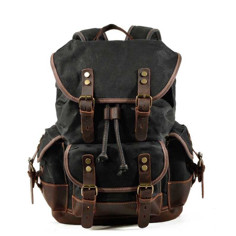 Puede incluir: Una mochila de lona negra con detalles y hebillas de cuero marr&oacute;n. La mochila tiene un cierre de cord&oacute;n y varios bolsillos.