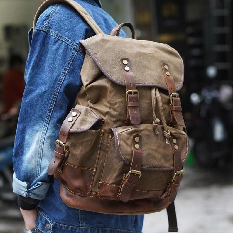 Puede incluir: Una mochila de lona marr&oacute;n con detalles de cuero y hebillas. La mochila tiene un compartimento principal grande, dos bolsillos delanteros y correas ajustables.