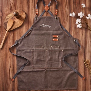 May include: A brown canvas apron with adjustable straps and a large pocket. The apron has a leather detail on the straps and two leather tabs on the front. The apron is personalised with the name "Jimmy" embroidered in white.