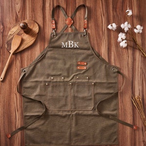 May include: A green canvas apron with brown leather straps and a large pocket. The apron has a white embroidered monogram "MBK" on the front.