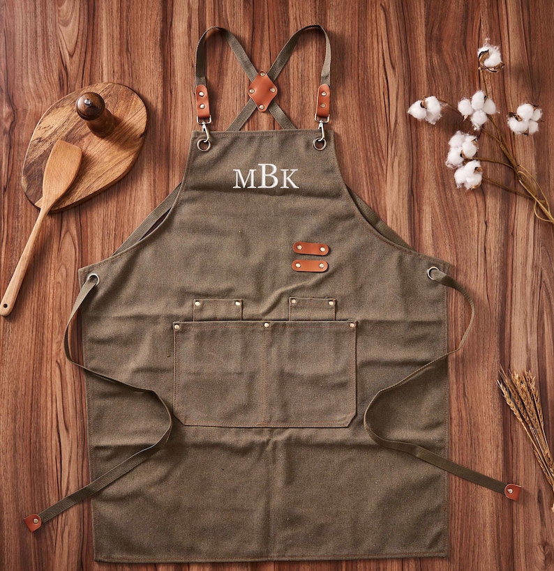 May include: A green canvas apron with brown leather straps and a large pocket. The apron has a white embroidered monogram "MBK" on the front.