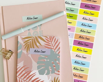 May include: Two pink notebooks with tropical leaf designs and the name "Melissa Connor" written on them. The notebooks are on a white surface with a set of colorful labels with the name "Melissa Connor" written on them.