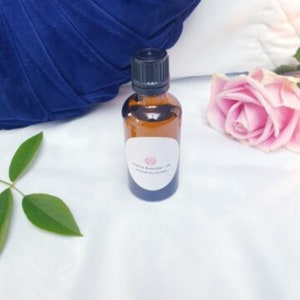 May include: A small amber glass bottle with a black cap, filled with a clear liquid. The bottle has a white label with the text "Luxury Blends" and a pink heart logo. The bottle is sitting on a white surface with a blue pillow and a pink rose in the background.