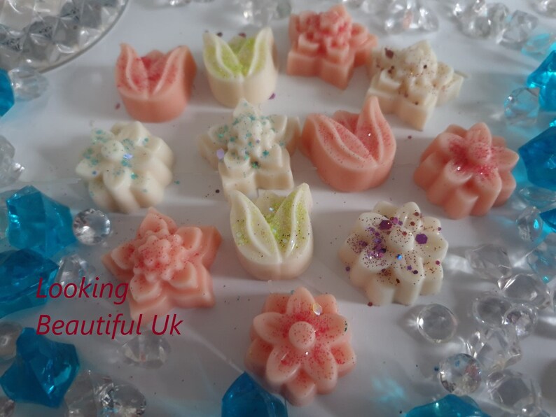 May include: Assortment of 12 small wax melts in various flower shapes. The melts are in shades of pink, white, and green with glitter accents.