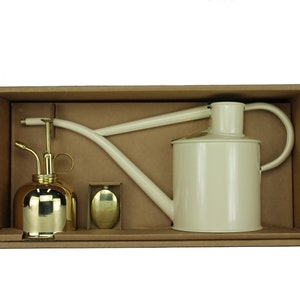 Könnte beinhalten: Ein Set aus zwei Gießkannen in einem Karton. Die größere Kanne ist weiß mit einem Messing-Ausguss und -Griff. Die kleinere Kanne ist golden mit einem Messing-Ausguss und -Griff.