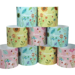 Może przedstawiać: Stos cylindrycznych pojemników z kwiatowymi wzorami i nazwami ziół. Pojemniki przedstawiają kolorowe akwarelowe ilustracje kwiatów, motyli i liści. Nazwy ziół są napisane odręcznie.