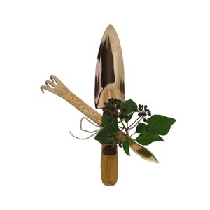Könnte beinhalten: Ein Set aus zwei Gartengeräten mit Holzgriffen. Die Geräte sind eine Schaufel und eine Handharke. Die Geräte sind mit Schnur zusammengebunden und mit einem Zweig grüner Blätter und kleinen braunen Beeren verziert.