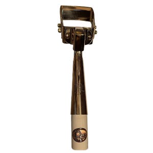 May include: A gold-toned metal tufting tool featuring a rectangular head with visible screws and a tapered neck. The handle is made of light-colored wood and displays a circular logo with the text PKS and a graphic of a spinning wheel.