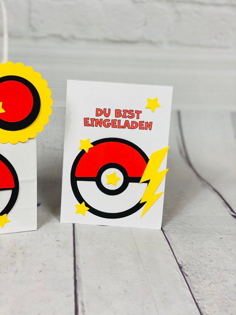 Puede incluir: Una tarjeta blanca con el texto "DU BIST EINGELADEN" en negro. La tarjeta presenta una Pok&eacute;bola roja y blanca con un rayo amarillo y estrellas amarillas.