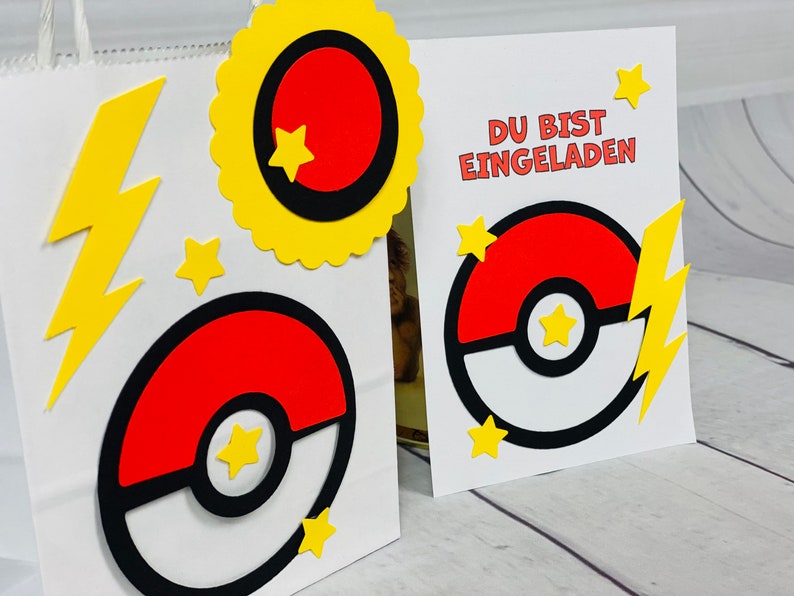 Puede incluir: Una bolsa de papel blanca con un rayo amarillo, una estrella amarilla y un dise&ntilde;o de Pok&eacute; Ball rojo y blanco. Una tarjeta blanca con el texto "DU BIST EINGELADEN" y un dise&ntilde;o de Pok&eacute; Ball rojo y blanco.