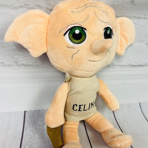 Könnte beinhalten: Ein Plüschtier von Dobby dem Hauself aus der Harry-Potter-Reihe. Das Spielzeug ist beige mit großen grünen Augen und einer kleinen, spitzen Nase. Es trägt eine beige Weste mit dem Wort "CELINA" bestickt auf der Vorderseite. Das Spielzeug sitzt auf einer Holzoberfläche.