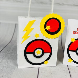 Puede incluir: Una bolsa de regalo de papel blanco con una Pok&eacute;bola roja y blanca, un rayo amarillo y una estrella amarilla. La bolsa tiene un asa amarilla.