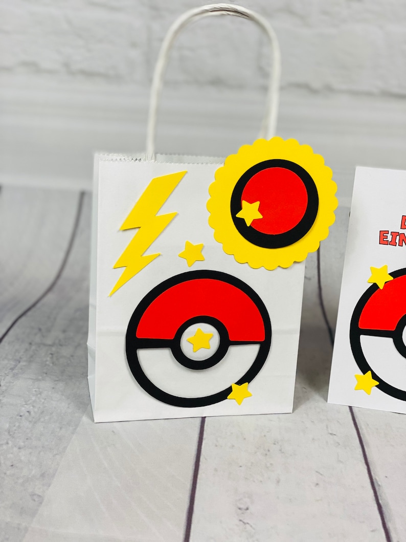 Puede incluir: Una bolsa de regalo de papel blanco con una Pok&eacute;bola roja y blanca, un rayo amarillo y una estrella amarilla. La bolsa tiene un asa amarilla.
