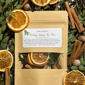 Könnte beinhalten: Eine braune Papiertüte mit einem durchsichtigen Fenster, das eine Mischung aus getrockneten Früchten, Gewürzen und Kräutern zeigt. Die Tüte ist mit "Holiday Simmer Pot Mix" beschriftet und enthält eine Zutatenliste: Cranberries, Muskatnuss, Nelken, Sternanis, Zimtstangen, Orangen und Kiefer. Die Tüte ist von Grünpflanzen und anderen natürlichen Elementen umgeben.