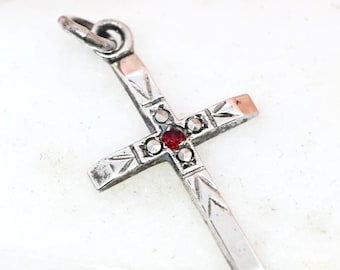 May include: A silver-toned cross pendant with a small red gemstone at the center. The cross features etched detailing and a loop for a chain. The pendant is displayed on a white surface with a soft pink background.