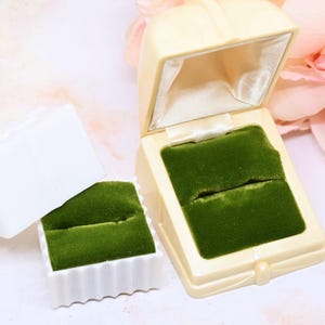 Puede incluir: Caja de anillos de color marfil con interior de terciopelo verde. Una caja más pequeña a juego con interior de terciopelo verde está al lado. Las cajas están sobre una superficie clara con un fondo floral rosa.