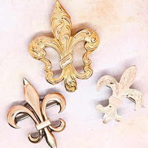 May include: Three gold-toned fleur-de-lis brooches. The brooches are ornate and detailed, with intricate designs.