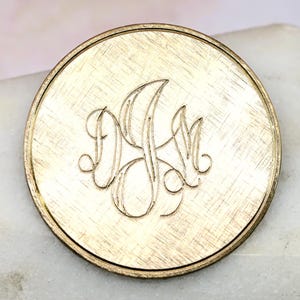 Peut inclure: Un disque rond en métal doré avec une finition brossée et un monogramme des lettres "D.K.M." gravé en écriture cursive.