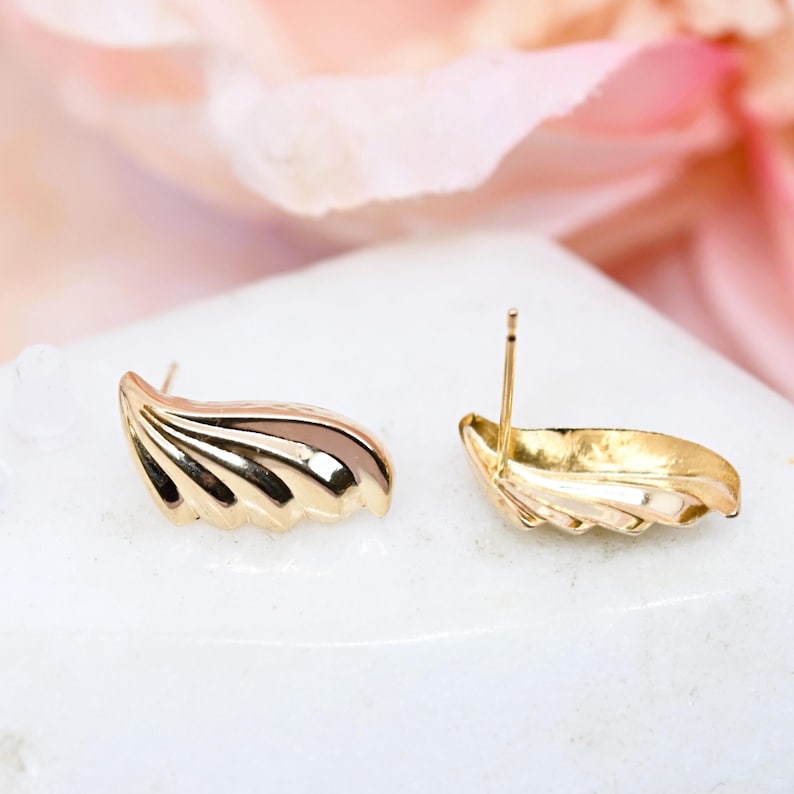 Puede incluir: Un par de pendientes de bot&oacute;n en forma de concha de color dorado. Los pendientes est&aacute;n hechos de un metal liso y brillante y tienen una forma ligeramente curvada.