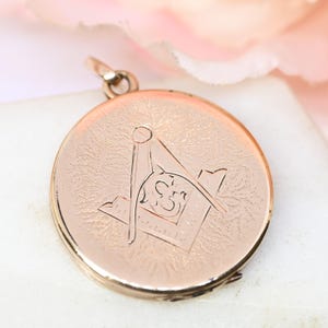 Peut inclure: Un pendentif rond de couleur or rose avec un symbole maçonnique gravé sur le devant. Le pendentif a un fond texturé et une petite boucle en haut pour une chaîne. Le symbole maçonnique comprend un carré, un compas et la lettre "G".