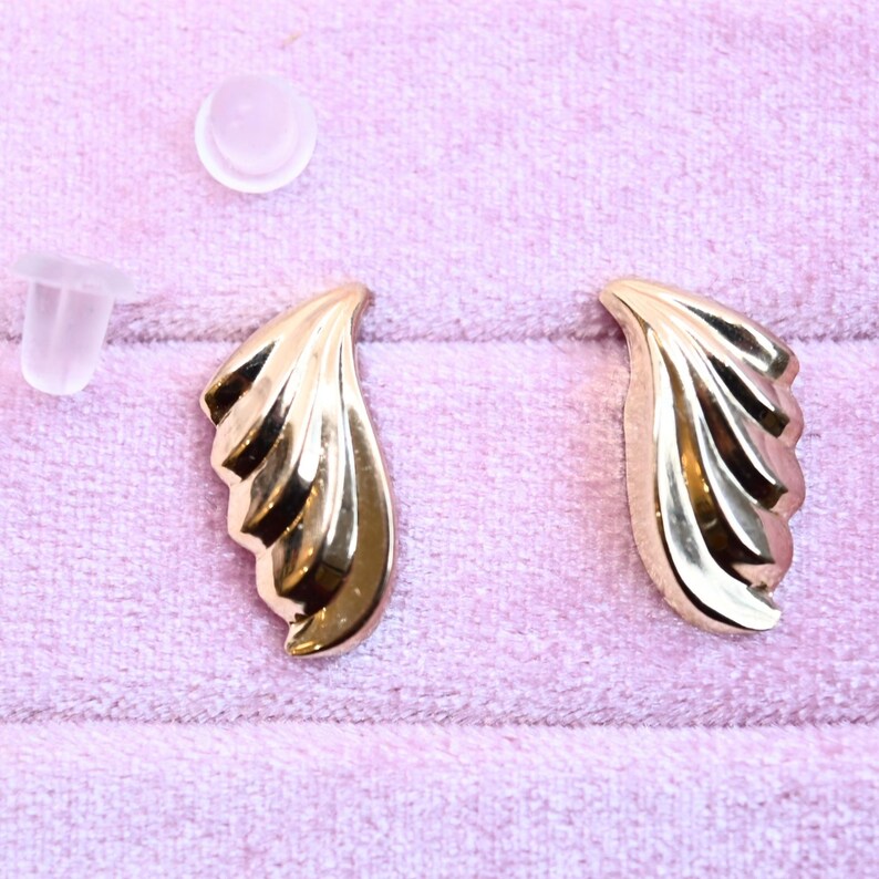 Puede incluir: Un par de pendientes de color dorado con un dise&ntilde;o en forma de abanico. Los pendientes est&aacute;n sobre una superficie de terciopelo rosa.