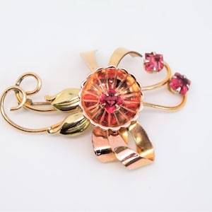 May include: A vintage gold-tone brooch with a floral design. The brooch features a central flower with a red gemstone center and surrounding petals. Two smaller red gemstones are set on curved stems. The design includes gold-tone leaves and ribbons.