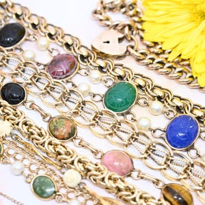 Puede incluir: Una colecci&oacute;n de pulseras de cadena de oro con diferentes amuletos de piedras preciosas de colores, incluyendo verde, azul, rosa y marr&oacute;n. Las pulseras est&aacute;n superpuestas sobre una superficie blanca.