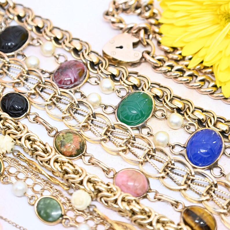 Puede incluir: Una colecci&oacute;n de pulseras de cadena de oro con diferentes amuletos de piedras preciosas de colores, incluyendo verde, azul, rosa y marr&oacute;n. Las pulseras est&aacute;n superpuestas sobre una superficie blanca.