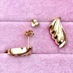 Puede incluir: Un par de pendientes de tono dorado con un dise&ntilde;o curvo en forma de onda. Los pendientes se muestran sobre una superficie de terciopelo rosa.