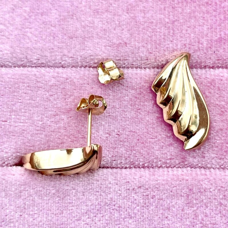 Puede incluir: Un par de pendientes de tono dorado con un dise&ntilde;o curvo en forma de onda. Los pendientes se muestran sobre una superficie de terciopelo rosa.