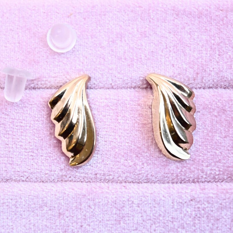 Puede incluir: Un par de pendientes de color dorado con un dise&ntilde;o en forma de abanico. Los pendientes est&aacute;n sobre una superficie de terciopelo rosa.