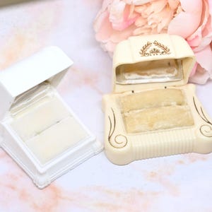 Pode incluir: Duas caixas de anel vintage. Uma é branca, retangular e aberta, revelando um interior creme. A outra é creme, com um topo curvo e detalhes dourados decorativos. Ambas as caixas têm inserções de veludo creme.