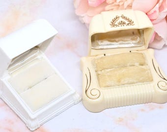 May include: Two vintage ring boxes. One is white, rectangular, and open, revealing a cream-colored interior. The other is off-white, with a curved top and decorative gold accents. Both boxes have cream-colored velvet inserts.