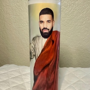 May include: A clear glass prayer candle featuring a portrait of a man with a beard, dressed in a robe and red cloth. The candle is cylindrical and has a white wax interior.