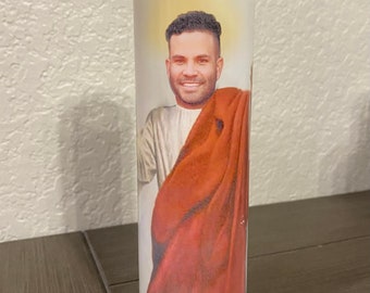 May include: A clear glass candle with a white label featuring a smiling person in a white robe and red shawl. The candle is cylindrical and stands upright on a dark surface.