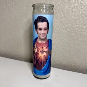 May include: A glass prayer candle featuring a portrait of a man with dark hair, dressed in blue robes, and a heart with a flame. The candle has a light blue background and is set on a white surface.