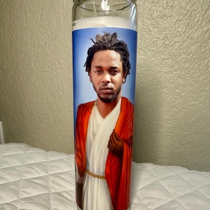 May include: A glass prayer candle featuring a portrait of a man in a red robe and white garment against a blue background. The candle has a white wax interior and a clear glass exterior.