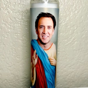 May include: A clear glass candle featuring a printed image of a smiling man in a religious pose. The man is wearing a blue and red robe. The candle is cylindrical and filled with white wax.