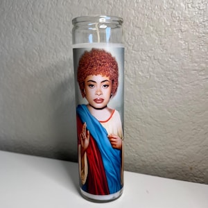 Peut inclure: Une bougie de prière en verre avec l'image d'une personne aux cheveux bouclés roux, vêtue d'une robe blanche et bleue. La bougie a une base en cire blanche et une mèche.