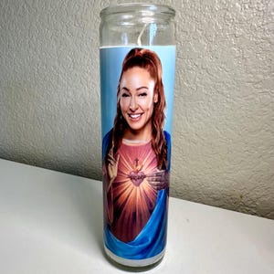 May include: A clear glass candle with a printed image of a woman with auburn hair and a blue background. The woman is wearing a pink and red top with a heart design. The candle has a white wick.