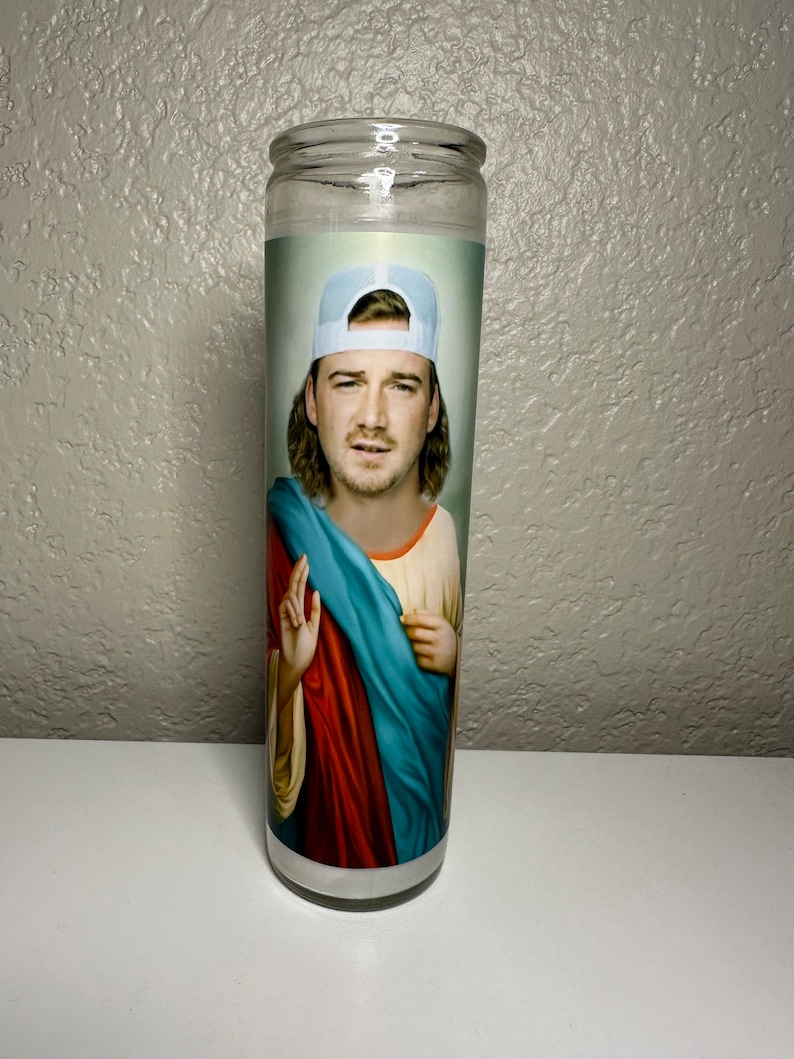Puede incluir: Una vela de oraci&oacute;n de vidrio transparente con la imagen de un hombre con una gorra de b&eacute;isbol azul claro. La imagen del hombre est&aacute; superpuesta a la vestimenta de una figura religiosa, con una t&uacute;nica roja y azul. La vela es cil&iacute;ndrica y est&aacute; sobre una superficie blanca.