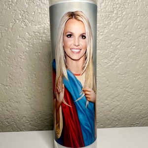 May include: A clear glass prayer candle with a portrait of a blonde woman with long hair, wearing a blue and red garment. The candle is cylindrical and filled with white wax.