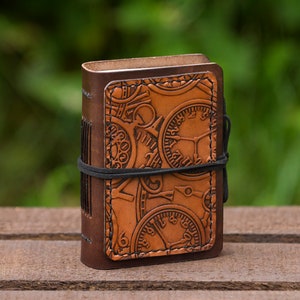Può includere: Un piccolo diario in pelle marrone con un design steampunk con quadranti dell'orologio e ingranaggi in rilievo. Il diario è chiuso e ha un elastico nero avvolto intorno.
