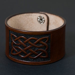May include: A brown leather bracelet with a Celtic knot design embossed on the front. The bracelet has a silver snap closure.