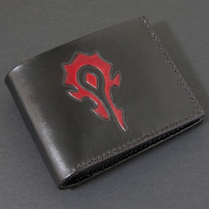 Könnte beinhalten: Schwarze Ledergeldbörse mit rotem Emblem. Die zweifach gefaltete Geldbörse hat einen genähten Rand und ein rotes Symbol auf der Vorderseite. Die Geldbörse ist dunkel und scheint aus Leder zu sein.