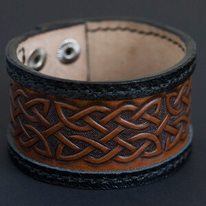 May include: A brown leather bracelet with a Celtic knot design. The bracelet has a black leather trim and two silver snaps.
