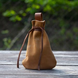 以下が含まれることがあります： 巾着型の茶色の革製ポーチ。紐で開閉し、ループ状の持ち手が付いています。ポーチは柔らかい革製で、底は丸みを帯びています。紐と持ち手は濃い茶色です。木製の表面に置かれています。
